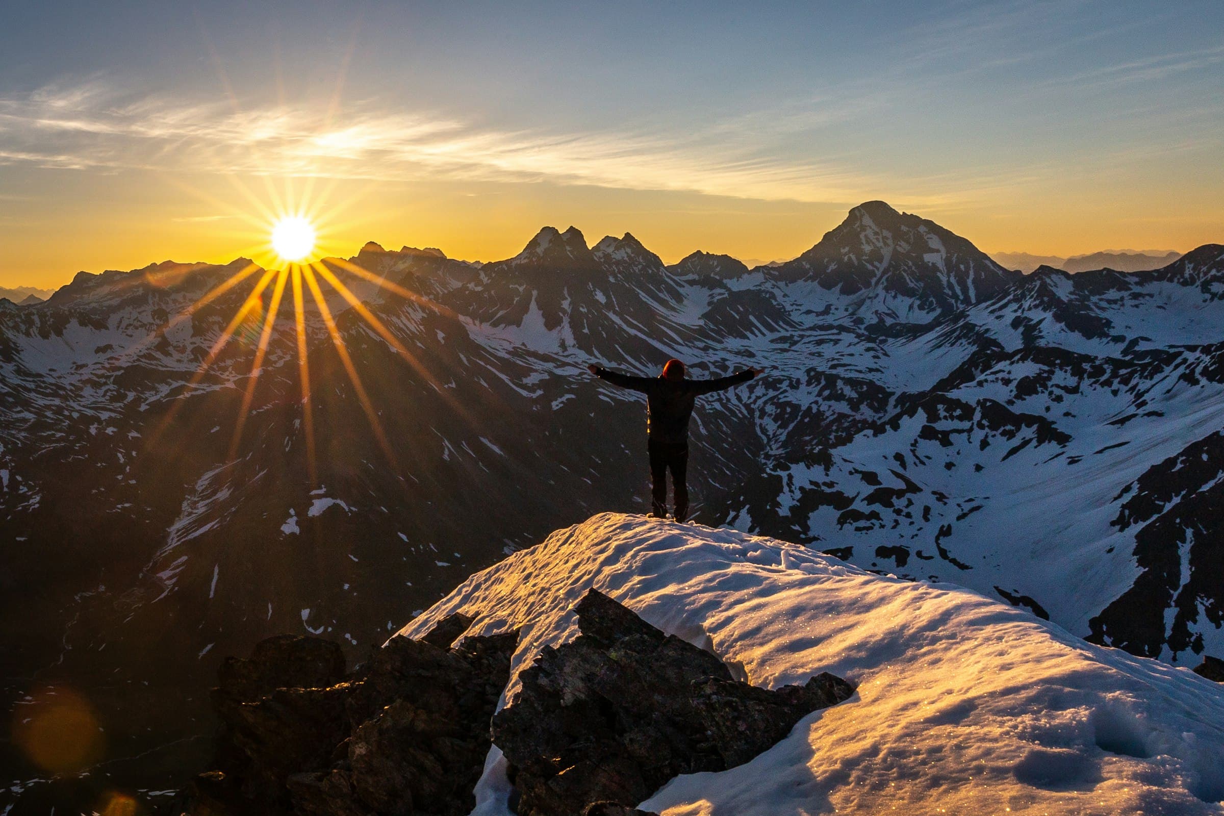 Hiker on a snowy summit with arms raised as the sun rises over the peaks